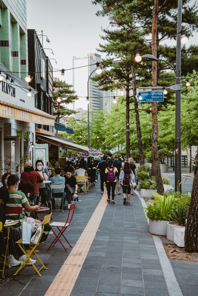 Well-lit daytime street in Seoul with people walking and sitting at streetside cafes and restaurants.